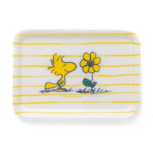 Könnte beinhalten: Ein weißes rechteckiges Tablett mit gelben Streifen und einem Comicbild von Woodstock aus Peanuts, einem gelben Vogel, der eine gelbe Blume betrachtet. Der Text "© 2023 PEANUTS" ist auf dem Tablett aufgedruckt.