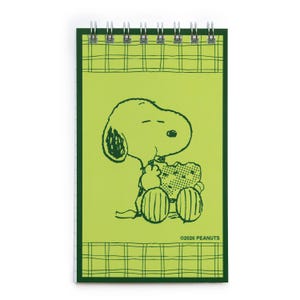 Pode incluir: Um bloco de notas com espiral, com uma capa verde limão e uma borda xadrez verde. A capa exibe uma ilustração em preto e branco de Snoopy de Peanuts, segurando um lanche. O texto "©2026 PEANUTS" está impresso na parte inferior.