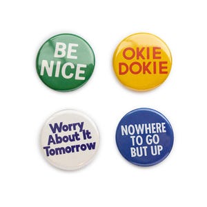 May include: Four round button pins with different sayings. The first pin is green with white text that says "BE NICE". The second pin is yellow with red text that says "OKIE DOKIE". The third pin is white with purple text that says "Worry About It Tomorrow". The fourth pin is blue with white text that says "NOWHERE TO GO BUT UP".