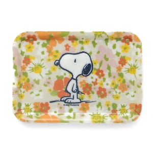 May include: A small, rectangular tray with a floral pattern in shades of orange, yellow, and white. The tray features a black and white illustration of Snoopy standing on a white background. The text "© 2023 PEANUTS" is printed in black at the bottom of the tray.