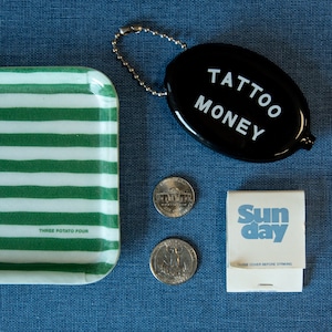K&ouml;nnte beinhalten: Eine gr&uuml;n-wei&szlig; gestreifte Keramik-Schale mit dem Text "Three Potato Four" auf der Unterseite. Eine schwarze Geldtasche mit einer silbernen Kette und dem Text "Tattoo Money" auf der Vorderseite. Zwei silberne 20-Cent-M&uuml;nzen und ein Streichholzschachtel mit dem Text "Sunday" in blauen Buchstaben und "Close cover before striking" in schwarzen Buchstaben.