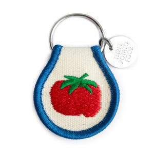 May include: A fabric keychain with a red tomato embroidered on a white background, outlined in blue. The keychain has a silver ring and a small silver tag that reads "THREE POTATO FOUR".