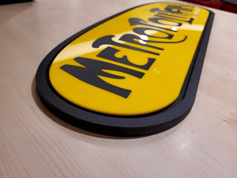 K&ouml;nnte beinhalten: Ein gelbes ovales Schild mit schwarzem Rand und dem Text "METROPOLITAN" in schwarzen Buchstaben.