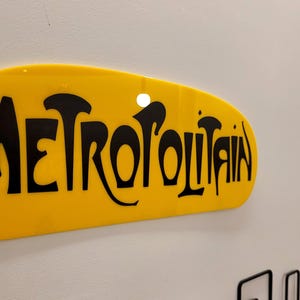 K&ouml;nnte beinhalten: Ein gelbes ovales Schild mit dem Wort "METROPOLITAIN" in schwarzen, stilisierten Buchstaben.