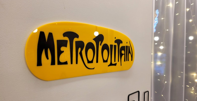 K&ouml;nnte beinhalten: Ein gelbes ovales Schild mit dem Wort "METROPOLITAIN" in schwarzen, stilisierten Buchstaben.