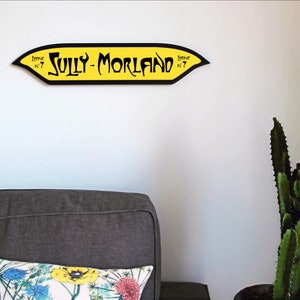 May include: A yellow and black sign with the text "Sully-Morland" in a decorative font. The sign is hanging on a white wall.