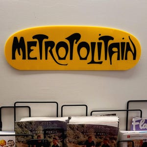 K&ouml;nnte beinhalten: Ein gelbes ovales Schild mit der Aufschrift "METROPOLITAIN" in schwarzen Buchstaben.