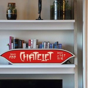 Peut inclure: Une enseigne rouge et blanche avec le texte "CHÂTELET" dans une police vintage, affichée sur une étagère blanche. L'enseigne comprend également le texte "LIGNES ABD" et "1-4-7 11-14". L'étagère contient des livres, des petites plantes et des objets décoratifs.