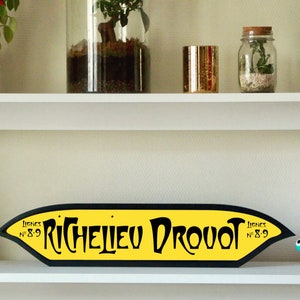 May include: A yellow and black sign with the text "Lignes N° 8-9 Richelieu Drouot Lignes N° 8-9" on a white shelf with plants and books.