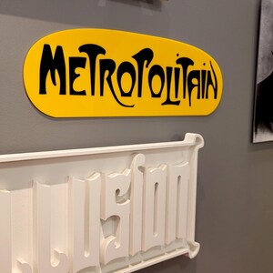 K&ouml;nnte beinhalten: Ein gelbes ovales Schild mit dem Wort "METROPOLITAIN" in schwarzen Buchstaben. Unter dem Schild befindet sich ein wei&szlig;es Schild mit dem Wort "USINE" in erhabenen Buchstaben.