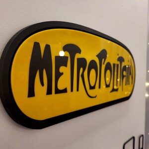 K&ouml;nnte beinhalten: Ein gelbes ovales Schild mit schwarzem Rand und dem Wort "METROPOLITAN" in schwarzen, stilisierten Buchstaben.