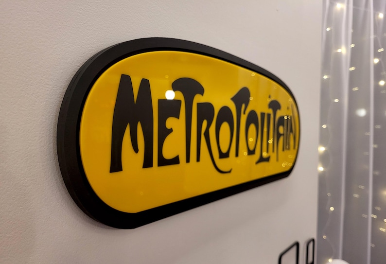 K&ouml;nnte beinhalten: Ein gelbes ovales Schild mit schwarzem Rand und dem Wort "METROPOLITAN" in schwarzen, stilisierten Buchstaben.