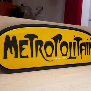 K&ouml;nnte beinhalten: Ein gelbes ovales Schild mit schwarzer Beschriftung, auf dem "METROPOLITAIN" steht. Das Schild befindet sich auf einer hellbraunen Holzoberfl&auml;che.