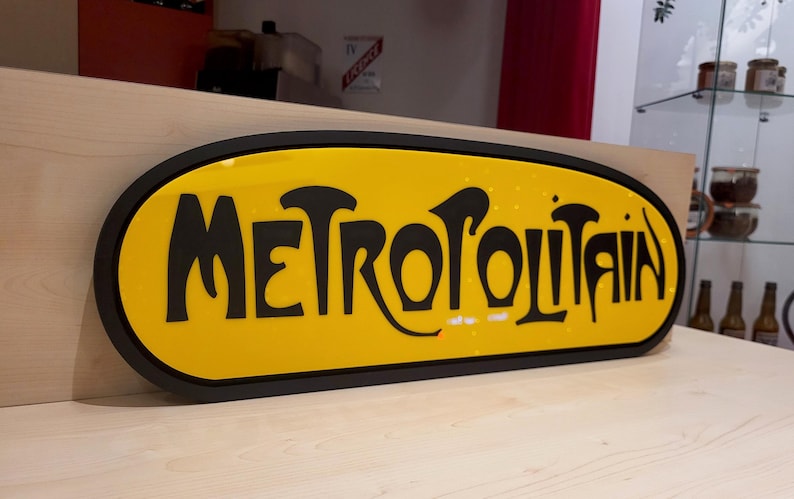 K&ouml;nnte beinhalten: Ein gelbes ovales Schild mit schwarzer Beschriftung, auf dem "METROPOLITAIN" steht. Das Schild befindet sich auf einer hellbraunen Holzoberfl&auml;che.