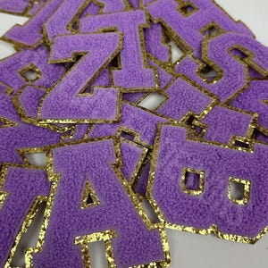 May include: Purple felt letter patches with gold glitter trim. The letters are in a varsity style font.