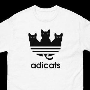 May include: White t-shirt with a black graphic of three black cats in a stylized Adidas logo. The text "adicats" is below the logo.