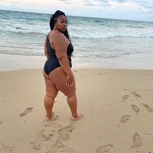 K&ouml;nnte beinhalten: Eine Person tr&auml;gt einen schwarzen Badeanzug mit orangefarbenen Akzenten und steht an einem Sandstrand mit dem Meer im Hintergrund.