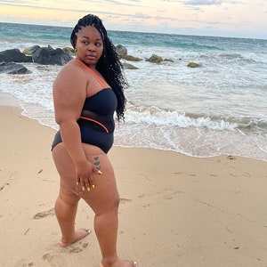 K&ouml;nnte beinhalten: Eine Frau in einem schwarzen Badeanzug mit orangefarbenen Akzenten steht an einem Sandstrand mit dem Meer im Hintergrund.