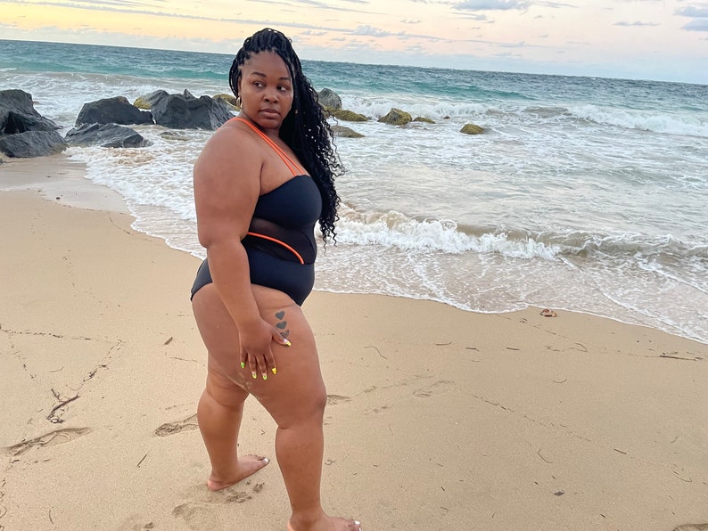 K&ouml;nnte beinhalten: Eine Frau in einem schwarzen Badeanzug mit orangefarbenen Akzenten steht an einem Sandstrand mit dem Meer im Hintergrund.