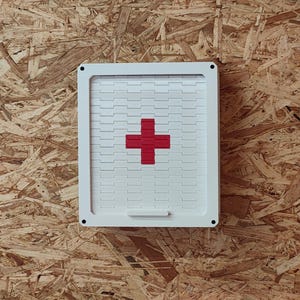 Può includere: Scatola di pronto soccorso bianca con una croce rossa sul davanti. La scatola è in plastica e ha un coperchio incernierato.