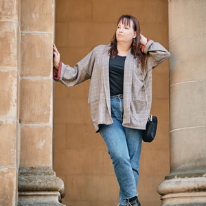 Pu&ograve; includere: Una donna indossa un blazer a quadri sopra una maglietta nera e jeans blu. &Egrave; in piedi tra due pilastri di pietra e ha una borsa nera sulla spalla.