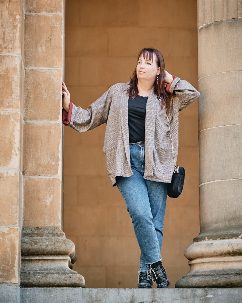 Pu&ograve; includere: Una donna indossa un blazer a quadri sopra una maglietta nera e jeans blu. &Egrave; in piedi tra due pilastri di pietra e ha una borsa nera sulla spalla.