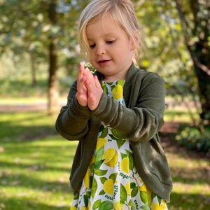 May include: A young girl wearing a green cardigan over a white dress with a lemon print. The dress has the text "best gift ever" repeated on it.