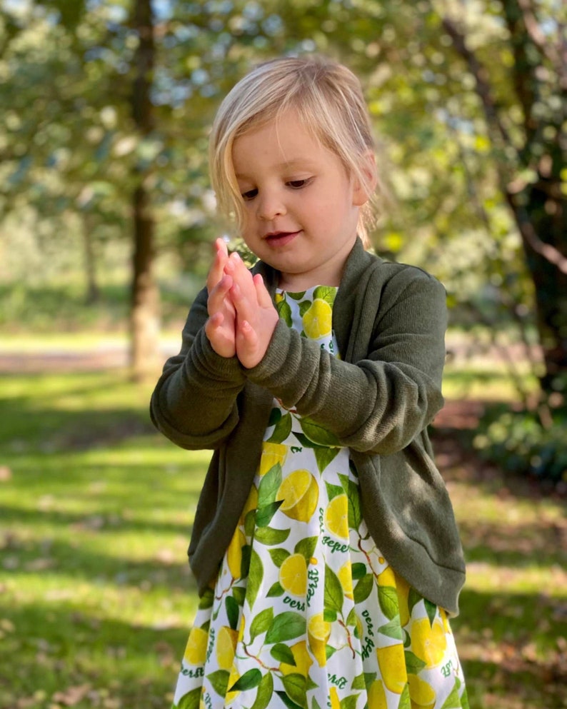 May include: A young girl wearing a green cardigan over a white dress with a lemon print. The dress has the text "best gift ever" repeated on it.