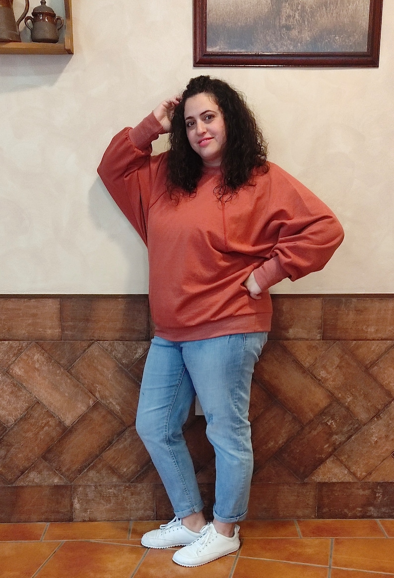 Pode incluir: Uma mulher vestindo uma camisa de moletom cor terracota de mangas compridas e caimento solto. Ela tamb&eacute;m est&aacute; vestindo jeans azul claro e t&ecirc;nis brancos.