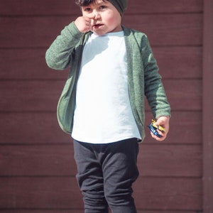 May include: A young boy wearing a green cardigan, a white t-shirt, black pants, and white sneakers with black stripes. He is wearing a green beanie and is holding a small toy car in his hand.