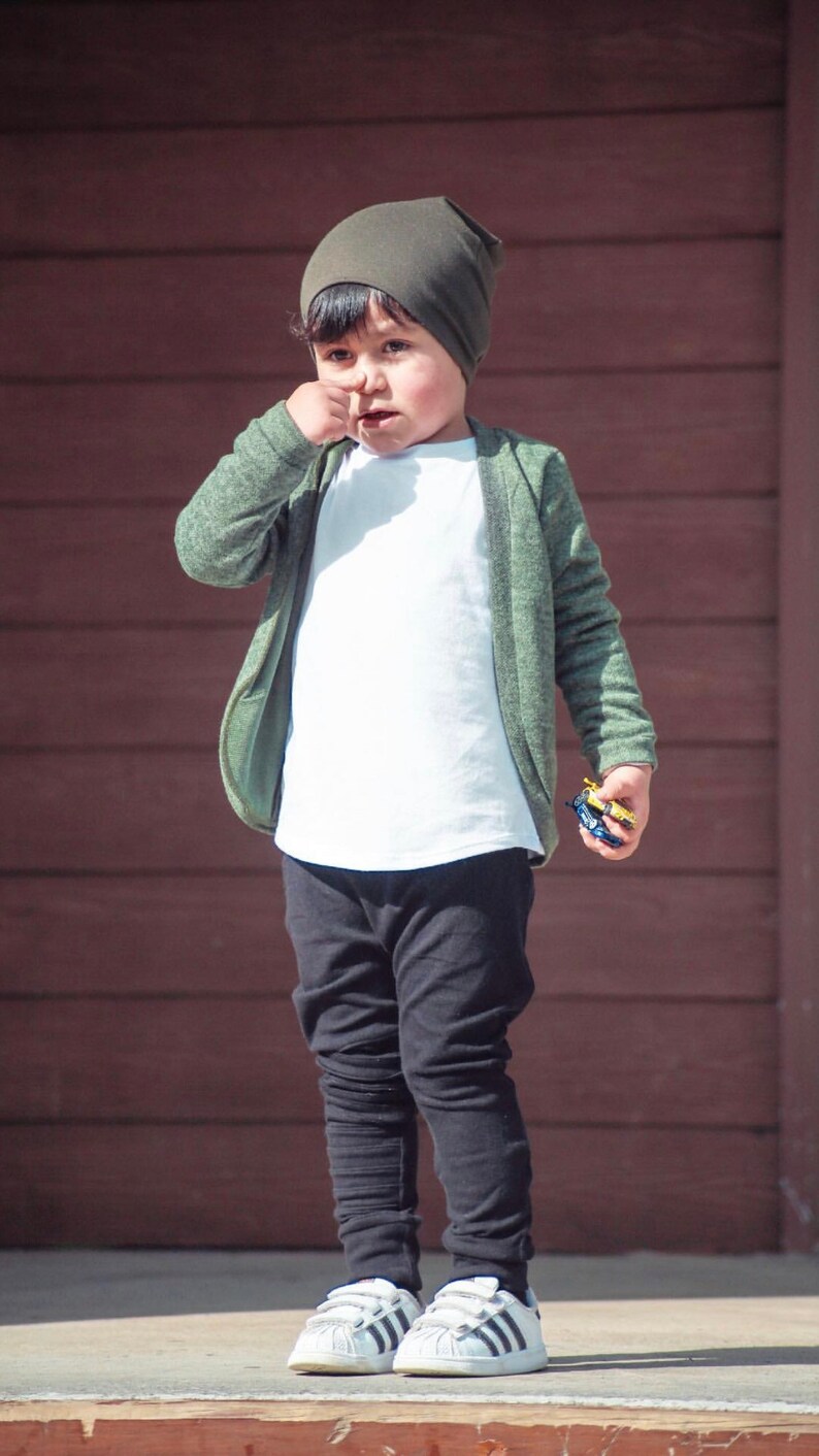 May include: A young boy wearing a green cardigan, a white t-shirt, black pants, and white sneakers with black stripes. He is wearing a green beanie and is holding a small toy car in his hand.