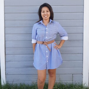 May include: A woman wearing a light blue and white striped shirt dress with a brown belt. She is standing in front of a grey wooden wall.