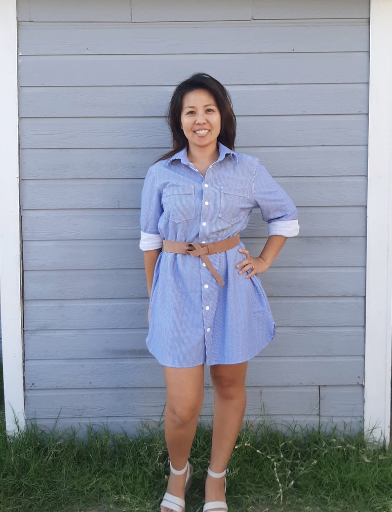 May include: A woman wearing a light blue and white striped shirt dress with a brown belt. She is standing in front of a grey wooden wall.