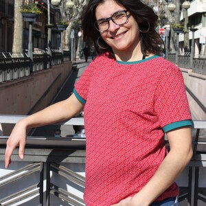 K&ouml;nnte beinhalten: Eine Frau tr&auml;gt ein rot-wei&szlig; gemustertes Shirt mit t&uuml;rkisfarbenem Besatz und blaue Jeans. Sie steht auf einer Br&uuml;cke und hat ihre Hand auf dem Gel&auml;nder.