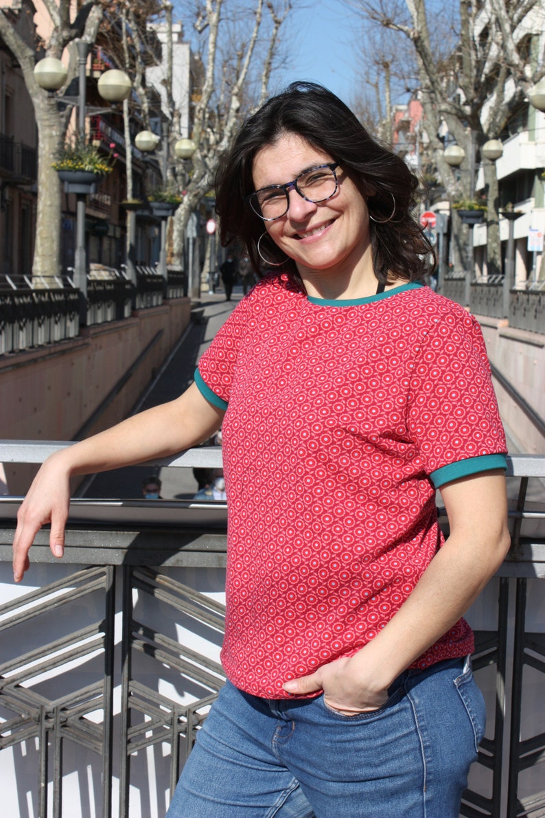 K&ouml;nnte beinhalten: Eine Frau tr&auml;gt ein rot-wei&szlig; gemustertes Shirt mit t&uuml;rkisfarbenem Besatz und blaue Jeans. Sie steht auf einer Br&uuml;cke und hat ihre Hand auf dem Gel&auml;nder.