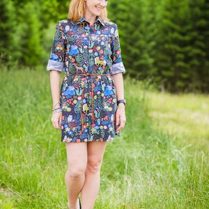 May include: A woman wearing a short-sleeved, dark blue dress with a colourful floral and animal print. The dress has a brown belt and a white collar. She is walking on a path in a grassy field.