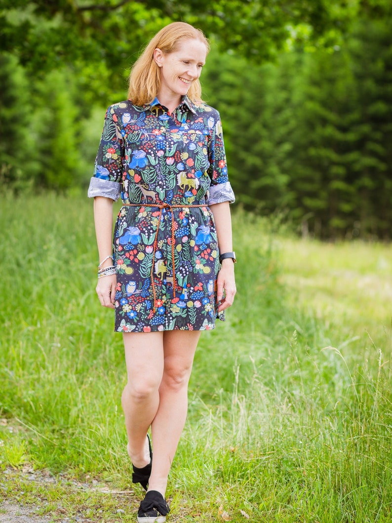 May include: A woman wearing a short-sleeved, dark blue dress with a colourful floral and animal print. The dress has a brown belt and a white collar. She is walking on a path in a grassy field.