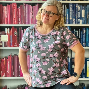 K&ouml;nnte beinhalten: Eine Frau tr&auml;gt ein graues T-Shirt mit einem pinkfarbenen Magnolienblumenmuster. Das Shirt hat einen pinkfarbenen Saum an Ausschnitt und &Auml;rmeln. Die Frau steht vor einem B&uuml;cherregal voller B&uuml;cher.