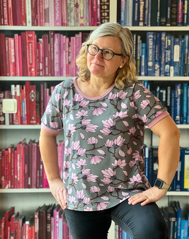 K&ouml;nnte beinhalten: Eine Frau tr&auml;gt ein graues T-Shirt mit einem pinkfarbenen Magnolienblumenmuster. Das Shirt hat einen pinkfarbenen Saum an Ausschnitt und &Auml;rmeln. Die Frau steht vor einem B&uuml;cherregal voller B&uuml;cher.