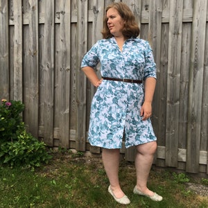 May include: A woman wearing a white and blue floral print shirt dress with a brown belt. The dress has a button-down front and three-quarter sleeves.