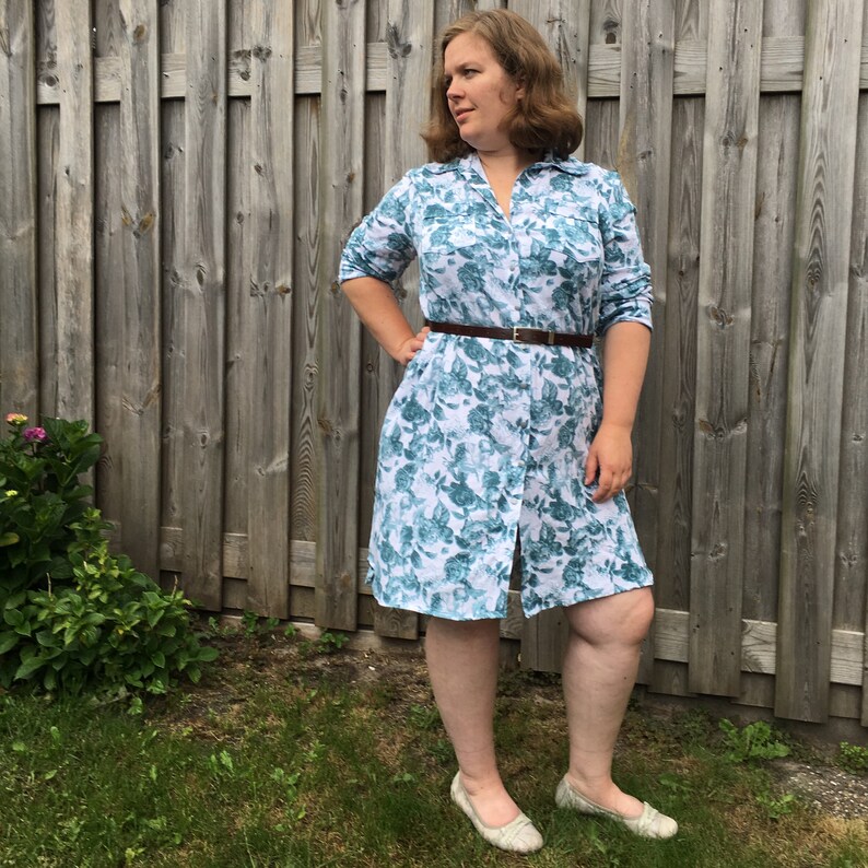 May include: A woman wearing a white and blue floral print shirt dress with a brown belt. The dress has a button-down front and three-quarter sleeves.