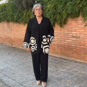 Pu&ograve; includere: Una donna indossa una giacca kimono nera e bianca con motivo geometrico a maniche lunghe e un completo pantalone nero.