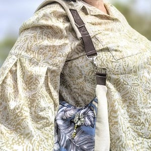Puede incluir: Un bolso de hombro con estampado floral azul y blanco con una correa de pana color crema y una correa de acento marr&oacute;n. El bolso tiene una flor decorativa y un adorno de cuentas. La persona lleva una camisa con botones estampada.