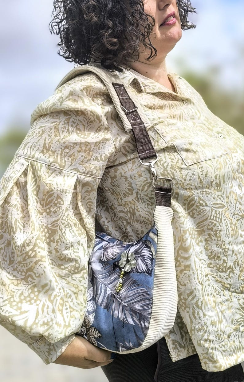 Puede incluir: Un bolso de hombro con estampado floral azul y blanco con una correa de pana color crema y una correa de acento marr&oacute;n. El bolso tiene una flor decorativa y un adorno de cuentas. La persona lleva una camisa con botones estampada.