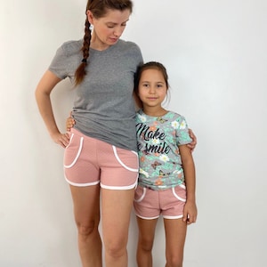 May include: Two people wearing pink and white athletic shorts. The adult is wearing a gray t-shirt and the child is wearing a light blue t-shirt with a floral pattern and the text "Make a Smile".