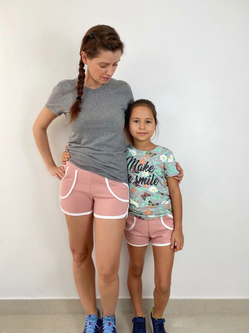May include: Two people wearing pink and white athletic shorts. The adult is wearing a gray t-shirt and the child is wearing a light blue t-shirt with a floral pattern and the text "Make a Smile".