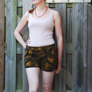 May include: A woman wearing a white tank top and black and gold patterned shorts. She is standing in front of a wooden fence.
