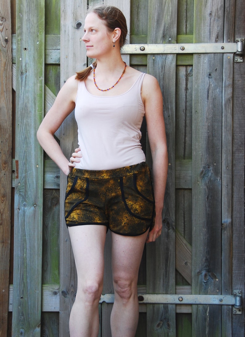 May include: A woman wearing a white tank top and black and gold patterned shorts. She is standing in front of a wooden fence.