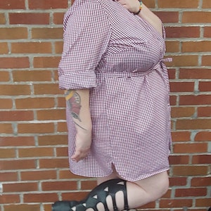 May include: A woman wearing a red and white gingham shirt dress with a red flower in her hair. She is wearing black gladiator sandals and is standing in front of a brick wall.