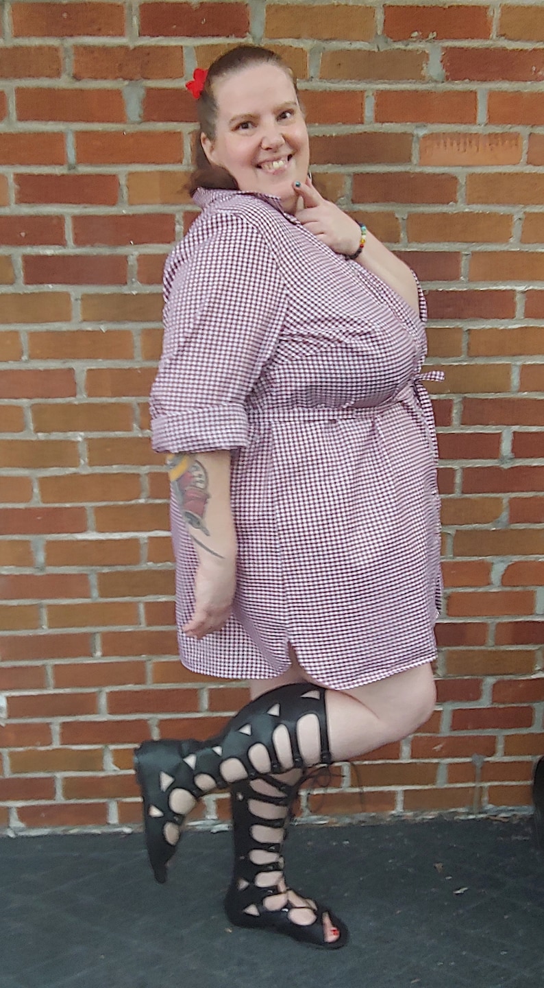 May include: A woman wearing a red and white gingham shirt dress with a red flower in her hair. She is wearing black gladiator sandals and is standing in front of a brick wall.
